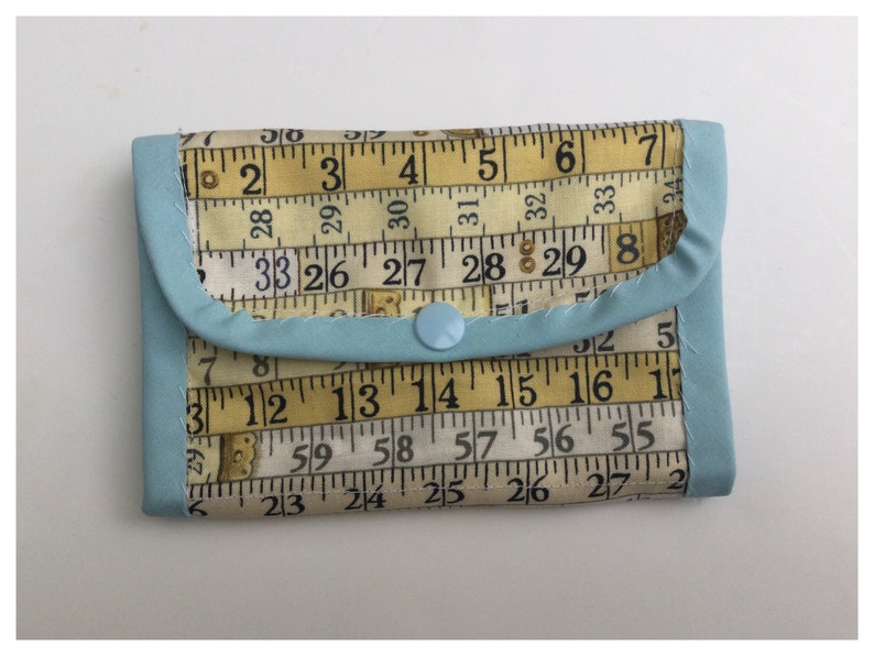 May include: A light blue fabric wallet with a snap closure. The wallet is made from fabric with a pattern of yellow measuring tapes with black numbers. The numbers on the tape measure include 2, 3, 4, 5, 6, 7, 8, 26, 27, 28, 29, 30, 31, 32, 33, 55, 56, 57, 58, 59, 12, 13, 14, 15, 16, 17, 23, 24, 25, 26, 27.