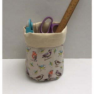 May include: A beige fabric storage bin with a cream coloured top. The bin is filled with various craft supplies, including a purple pair of scissors, a blue tape dispenser, and a wooden ruler. The bin is decorated with a pattern of colourful birds and leaves.