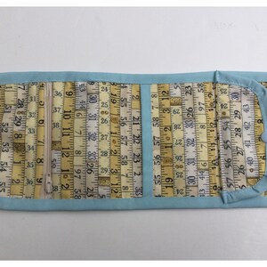 May include: A light blue fabric wallet with a zipper closure. The fabric is printed with a pattern of yellow measuring tapes with numbers. The wallet has a flap with a snap closure.