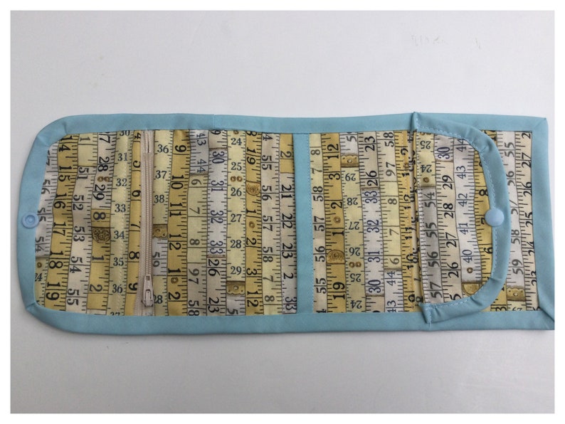 May include: A light blue fabric wallet with a zipper closure. The fabric is printed with a pattern of yellow measuring tapes with numbers. The wallet has a flap with a snap closure.
