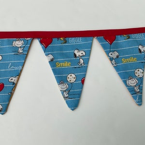 May include: A blue and white fabric banner with a red trim. The banner has a repeating pattern of Snoopy characters and the words "Giggle", "Smile", and "Laugh".