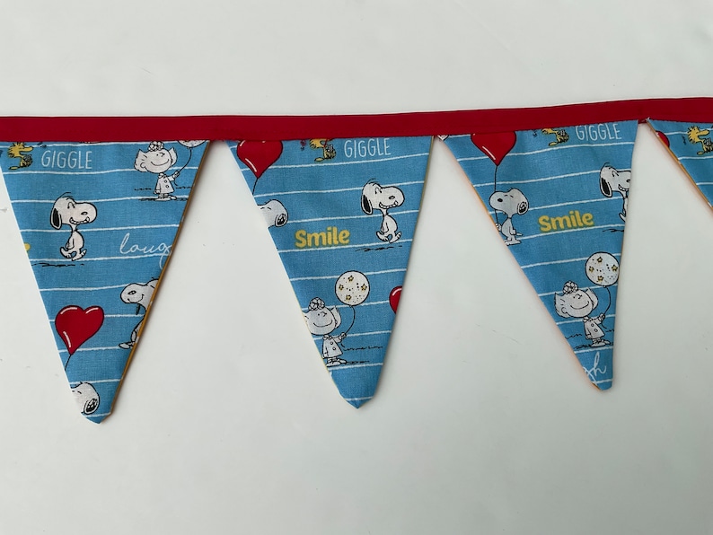 May include: A blue and white fabric banner with a red trim. The banner has a repeating pattern of Snoopy characters and the words "Giggle", "Smile", and "Laugh".