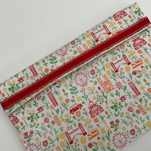 May include: A fabric pouch with a London-themed design. The white background is patterned with red, orange, and yellow illustrations of landmarks, flowers, and vehicles. A red ribbon with white stitching trims the top.