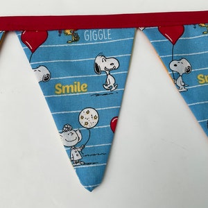 May include: A blue and white fabric banner with a red trim. The banner has a repeating pattern of Snoopy characters and the words "Giggle" and "Smile".