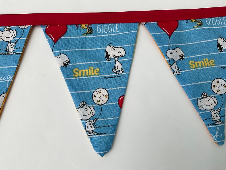 May include: A blue and white fabric banner with a red trim. The banner has a repeating pattern of Snoopy characters and the words "Giggle" and "Smile".