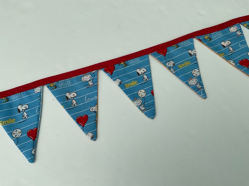 May include: A blue and white fabric banner with red trim. The banner has a repeating pattern of Snoopy characters and the words "Giggle" and "Smile".