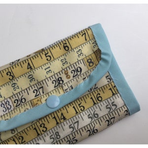 May include: A small blue fabric wallet with a yellow and white fabric design featuring a repeating pattern of a measuring tape with numbers. The wallet has a blue button closure.