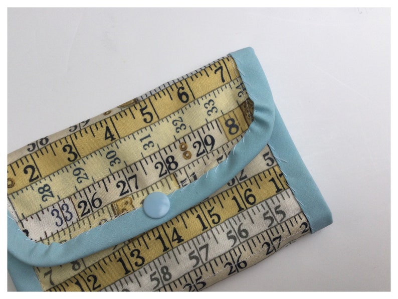 May include: A small blue fabric wallet with a yellow and white fabric design featuring a repeating pattern of a measuring tape with numbers. The wallet has a blue button closure.