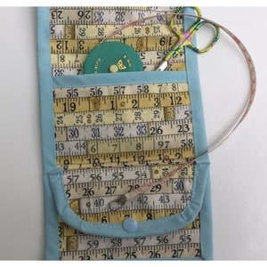 May include: A light blue fabric pouch with a flap closure. The pouch is decorated with a pattern of vintage measuring tapes. The pouch has a small pocket on the front and a button closure. A small green measuring tape and a pair of pink and green scissors are visible inside the pouch.