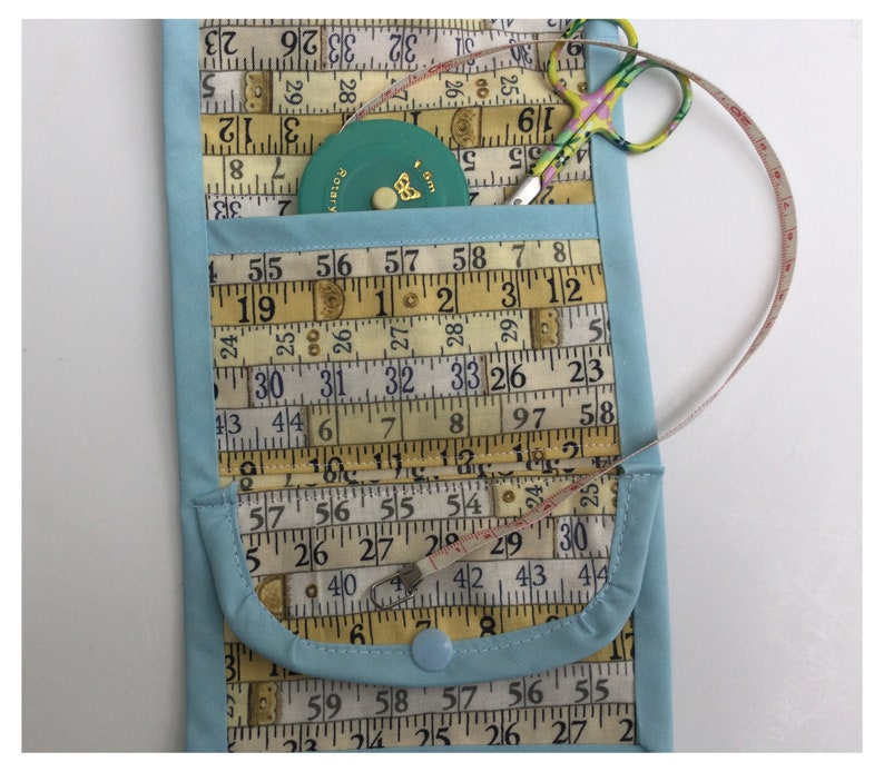 May include: A light blue fabric pouch with a flap closure. The pouch is decorated with a pattern of vintage measuring tapes. The pouch has a small pocket on the front and a button closure. A small green measuring tape and a pair of pink and green scissors are visible inside the pouch.