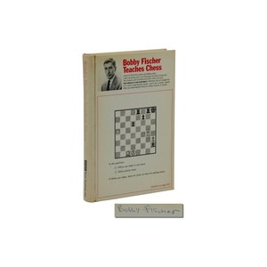 Può includere: Un libro bianco con una scacchiera in bianco e nero sulla copertina. Il libro è intitolato "Bobby Fischer Teaches Chess". La firma dell'autore, "Bobby Fischer", è scritta in inchiostro blu nella parte inferiore della copertina.