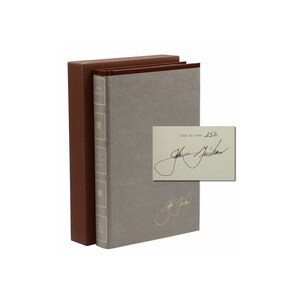 May include: A gray hardcover book with a brown slipcase. The book has a gold title on the spine that reads "The American Crisis". The book is signed by the author, "John Grisham", and includes the text "This is copy #252".