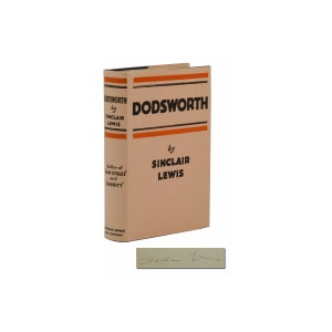 May include: A tan-colored book with an orange and black striped spine. The title "Dodsworth" is printed in black on the front cover, with the author's name "Sinclair Lewis" printed below. The book is signed by the author.