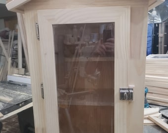 Puede incluir: Una pequeña biblioteca gratuita de madera sin terminar con una puerta de vidrio y un pestillo de metal. La biblioteca tiene un techo inclinado y está sentada sobre una superficie de madera.