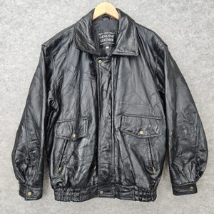 May include: A black leather jacket featuring a collared neckline, long sleeves with buttoned cuffs, and a front zipper closure. The jacket includes two front flap pockets with button accents and a ribbed hem. A label reads Genuine Leather.