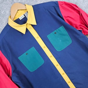 May include: A button-down shirt with a color-block design. The shirt features a navy blue body, yellow collar and button strip, green pockets, and red sleeves. The shirt is hanging on a wooden hanger.