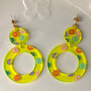 May include: A pair of yellow hoop earrings with a floral design. The earrings are made of acrylic and have a gold-tone finish.