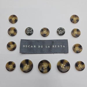 May include: A collection of buttons in various sizes and designs, featuring a tortoiseshell pattern in shades of brown and tan. Some buttons have a unique, textured appearance. A gray label with the text "OSCAR DE LA RENTA" is also present.