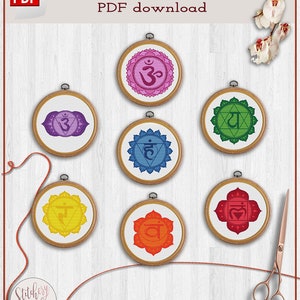 May include: Seven colourful embroidery hoops with cross stitch patterns of the seven chakras. The chakras are represented by symbols and colours. The hoops are arranged in a grid pattern on a white wooden background. The text "Cross stitch pattern PDF download" is at the top of the image.