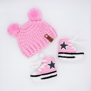 May include: Pink crocheted baby hat with pom poms and a brown tag that says "x stitch".  Two pink crocheted baby shoes with black and white stripes and a gray star.