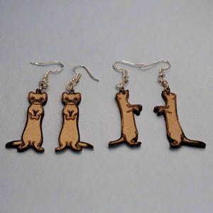 May include: A pair of dangle earrings featuring a brown wooden cutout of a ferret. The ferret is standing on its hind legs with its front paws raised. The earrings are made with silver hooks.