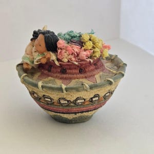May include: A decorative basket with a Native American theme. The basket is tan, green, and red with a woven design. It contains small figurines and colorful flowers. The figurines appear to be of Native American children.