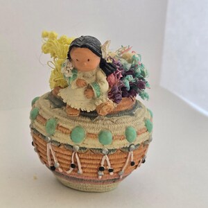 May include: A decorative figurine featuring a Native American girl sitting atop a woven basket. The basket is adorned with turquoise-colored accents and holds a colorful arrangement of dried flowers. The girl wears a white dress and has black hair.