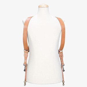 May include: A tan leather camera strap with adjustable straps and brass hardware. The strap is designed to be worn over the shoulders and across the chest.