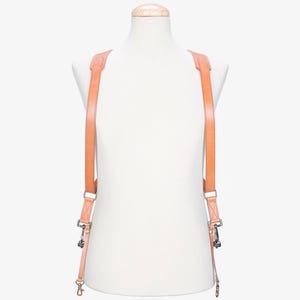 May include: A tan leather camera harness with silver-colored metal hardware. The harness is designed to be worn over the shoulders and has adjustable straps. The straps connect to the camera with metal clips.