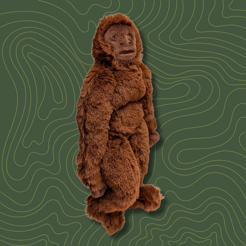 May include: A brown plush toy shaped like a Bigfoot with a furry texture. The toy has a human-like face with a wide mouth and large eyes.