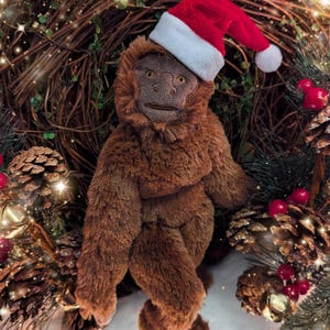 May include: A brown plush monkey wearing a red and white Santa hat. The monkey is sitting in a wreath of pine branches and red berries.