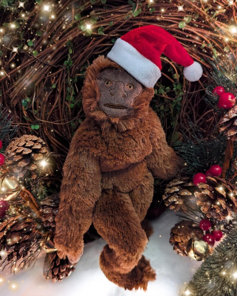 May include: A brown plush monkey wearing a red and white Santa hat. The monkey is sitting in a wreath of pine branches and red berries.