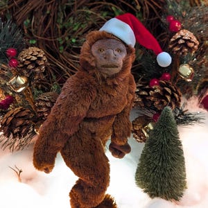 May include: A brown plush toy monkey wearing a red and white Santa hat. The monkey is standing on a white surface with a small green pine tree in the background.