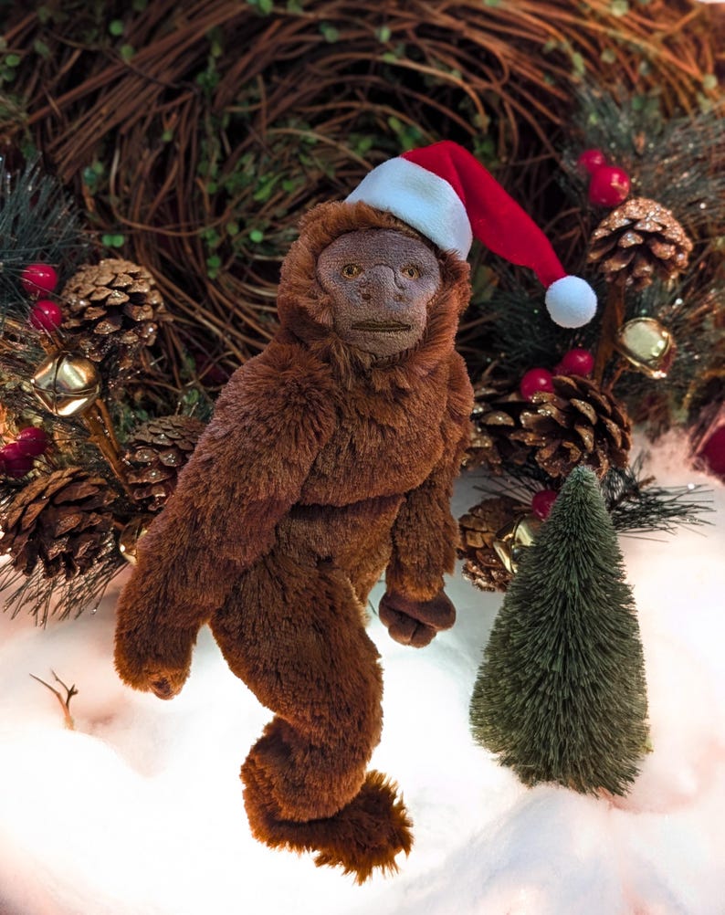 May include: A brown plush toy monkey wearing a red and white Santa hat. The monkey is standing on a white surface with a small green pine tree in the background.