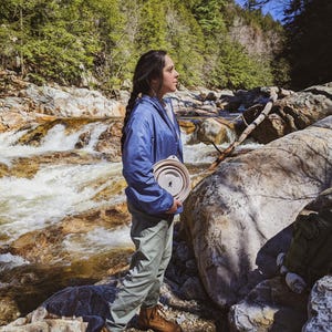 Peut inclure: Une personne portant une veste bleue et un pantalon vert tient un bol pliable de couleur claire avec une silhouette noire d'une personne. La personne se tient pr&egrave;s d'une rivi&egrave;re rocheuse avec une for&ecirc;t en arri&egrave;re-plan.