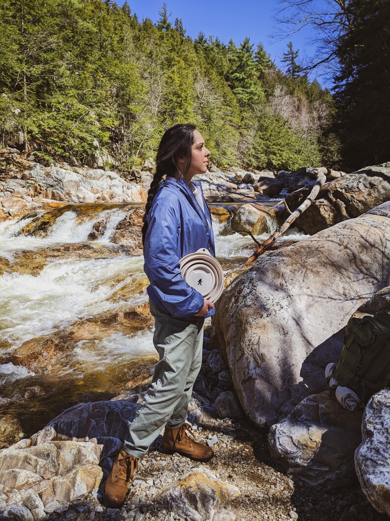 Peut inclure: Une personne portant une veste bleue et un pantalon vert tient un bol pliable de couleur claire avec une silhouette noire d'une personne. La personne se tient pr&egrave;s d'une rivi&egrave;re rocheuse avec une for&ecirc;t en arri&egrave;re-plan.