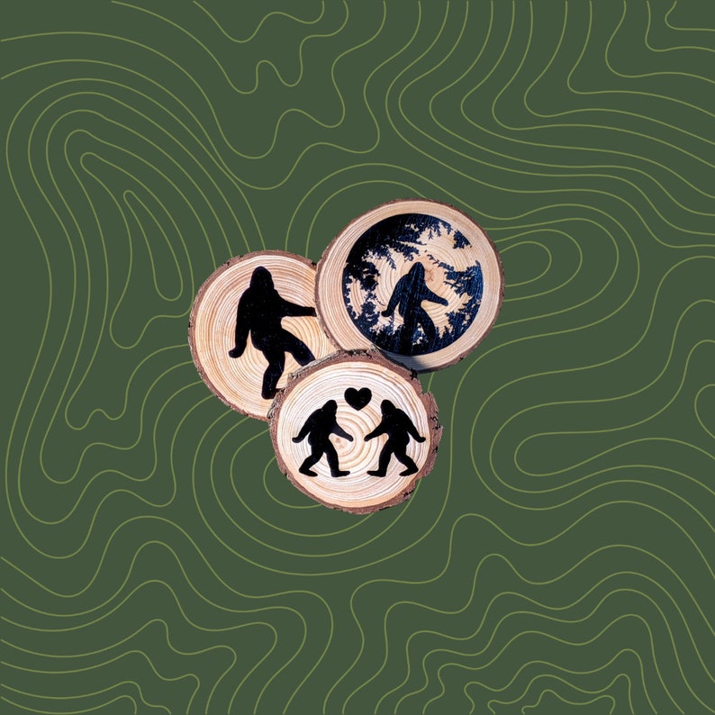 Pu&ograve; includere: Tre sottobicchieri in legno con un tema naturalistico. I sottobicchieri presentano una silhouette di un Bigfoot, una coppia di Bigfoots che si tengono per mano e una scena di bosco.