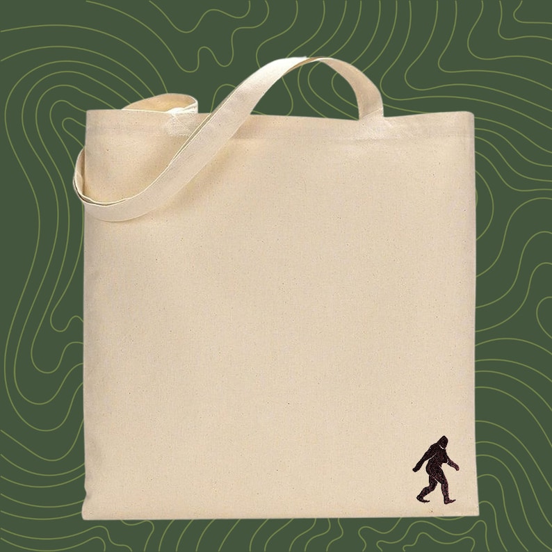 Op de afbeelding: Een natuurlijke canvas draagtas met een zwart silhouet van een Bigfoot die in de rechterbenedenhoek loopt.