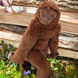 May include: A brown plush monkey toy with a long, furry tail. The monkey is standing on a bed of green moss and purple flowers.