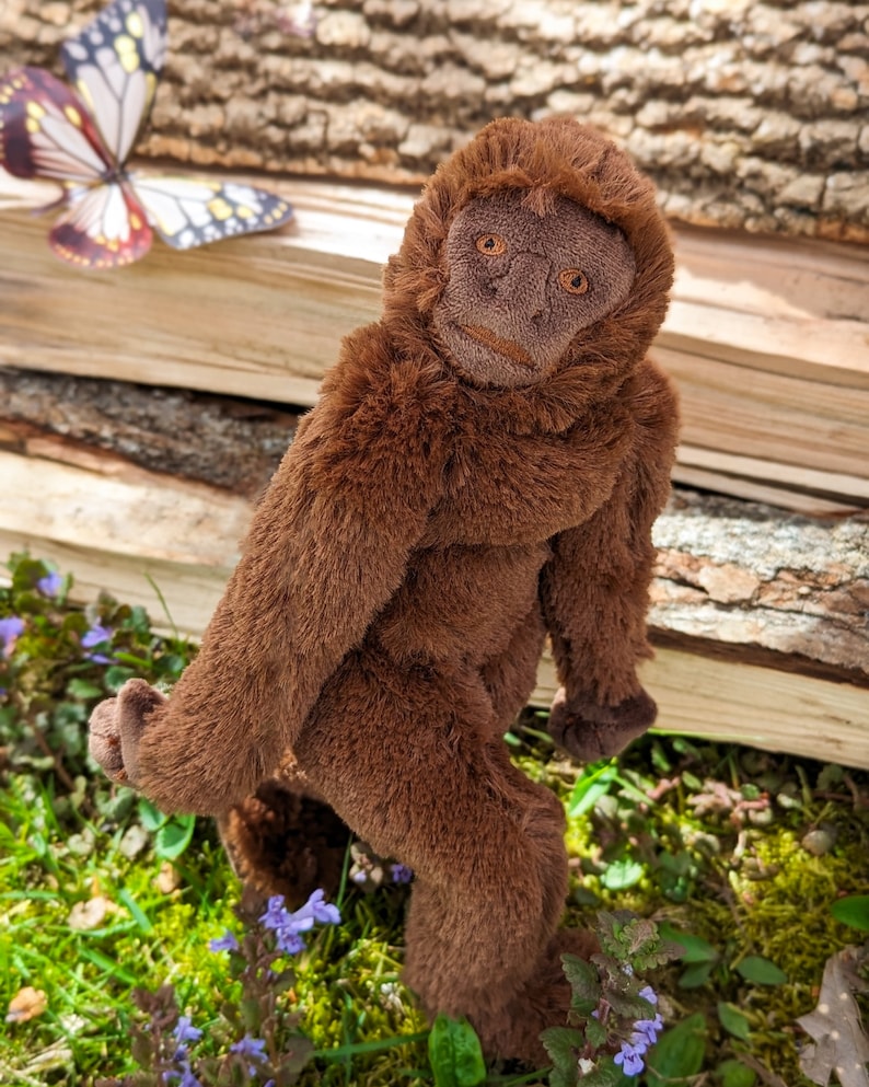May include: A brown plush monkey toy with a long, furry tail. The monkey is standing on a bed of green moss and purple flowers.