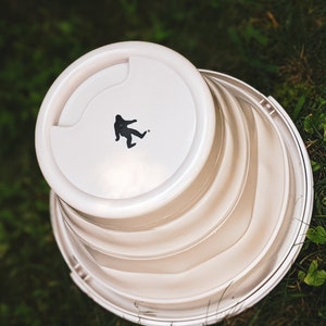 Peut inclure: Un seau pliable blanc avec une silhouette noire de Bigfoot sur le couvercle. Le seau est partiellement pli&eacute;, r&eacute;v&eacute;lant ses c&ocirc;t&eacute;s stri&eacute;s. L'image est prise d'en haut, sur fond d'herbe verte.