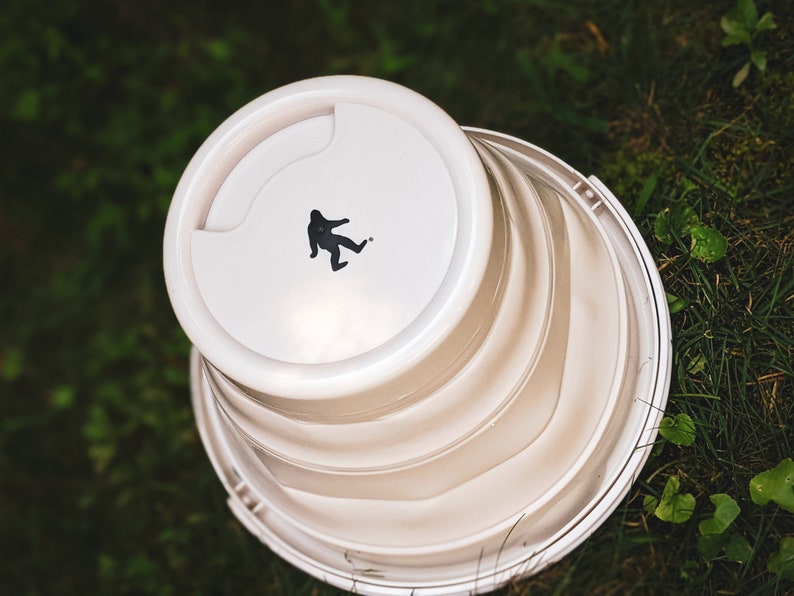 Peut inclure: Un seau pliable blanc avec une silhouette noire de Bigfoot sur le couvercle. Le seau est partiellement pli&eacute;, r&eacute;v&eacute;lant ses c&ocirc;t&eacute;s stri&eacute;s. L'image est prise d'en haut, sur fond d'herbe verte.