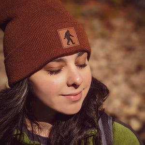 Pu&ograve; includere: Una donna indossa un berretto di lana marrone con una toppa in pelle che presenta la silhouette di un Bigfoot. Il berretto &egrave; tirato gi&ugrave; sulla fronte e lei guarda in basso con gli occhi chiusi. Indossa una giacca di pile verde.