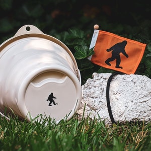 Peut inclure: Un seau beige avec une silhouette noire de Bigfoot, &agrave; c&ocirc;t&eacute; d'un petit drapeau orange avec un graphique de Bigfoot. Le drapeau est sur un petit poteau en bois. Une roche textur&eacute;e est &eacute;galement dans l'image.