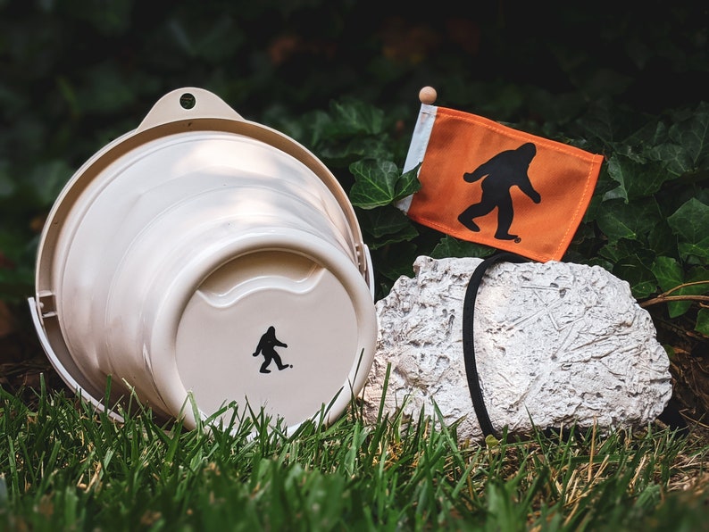 Peut inclure: Un seau beige avec une silhouette noire de Bigfoot, &agrave; c&ocirc;t&eacute; d'un petit drapeau orange avec un graphique de Bigfoot. Le drapeau est sur un petit poteau en bois. Une roche textur&eacute;e est &eacute;galement dans l'image.