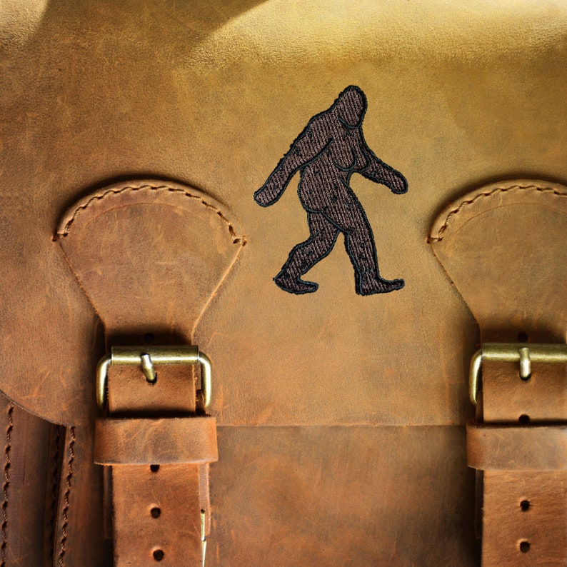 K&ouml;nnte beinhalten: Eine braune Ledertasche mit zwei Schnallen und einem schwarzen gestickten Bild von Bigfoot.