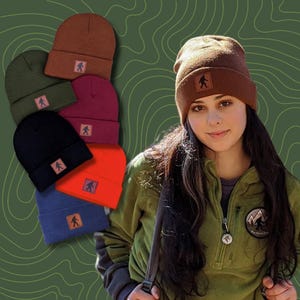 Könnte beinhalten: Ein Set aus sechs Strickmützen in verschiedenen Farben, jede mit einem braunen Lederpatch mit einer Bigfoot-Silhouette. Die Mützen sind in einem Stapel angeordnet, wobei die braune Mütze oben liegt. Eine Frau trägt eine braune Mütze und eine grüne Fleecejacke mit einem Patch einer Bergkette und einer Bigfoot-Silhouette.