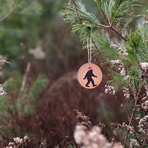 Op de afbeelding: Een houten kerstversiering met een silhouet van Bigfoot die een kerstmanmuts draagt. De versiering hangt aan een dennentak.