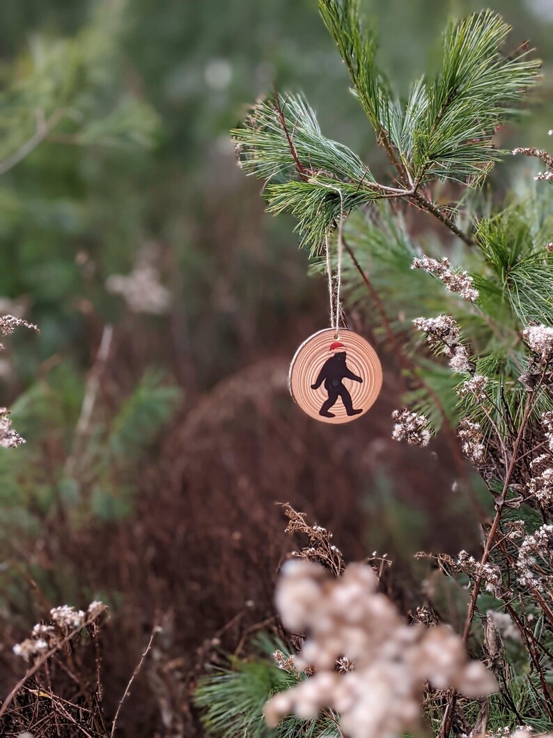 Op de afbeelding: Een houten kerstversiering met een silhouet van Bigfoot die een kerstmanmuts draagt. De versiering hangt aan een dennentak.