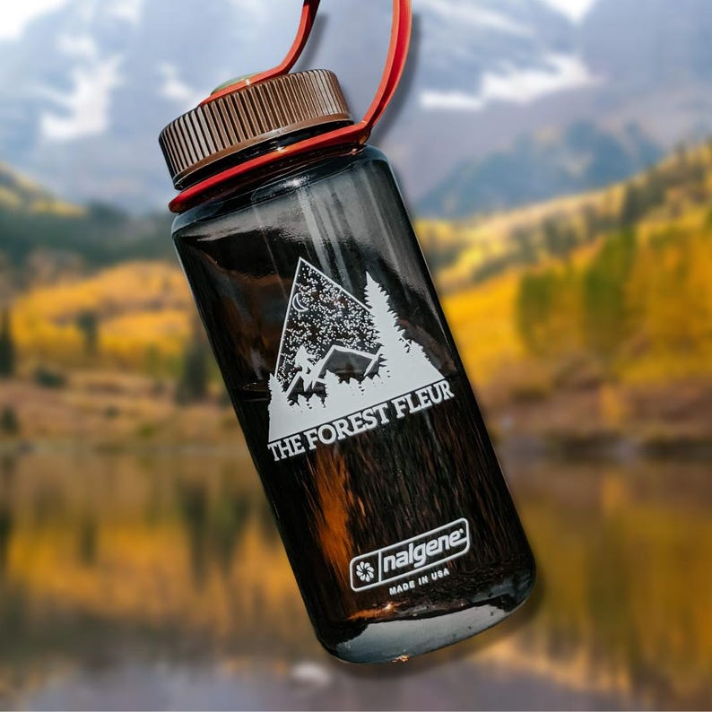 Może przedstawiać: Czarna butelka Nalgene z białym nadrukiem "The Forest Fleur" i grafiką księżyca i gwiazd. Butelka została wyprodukowana w USA.